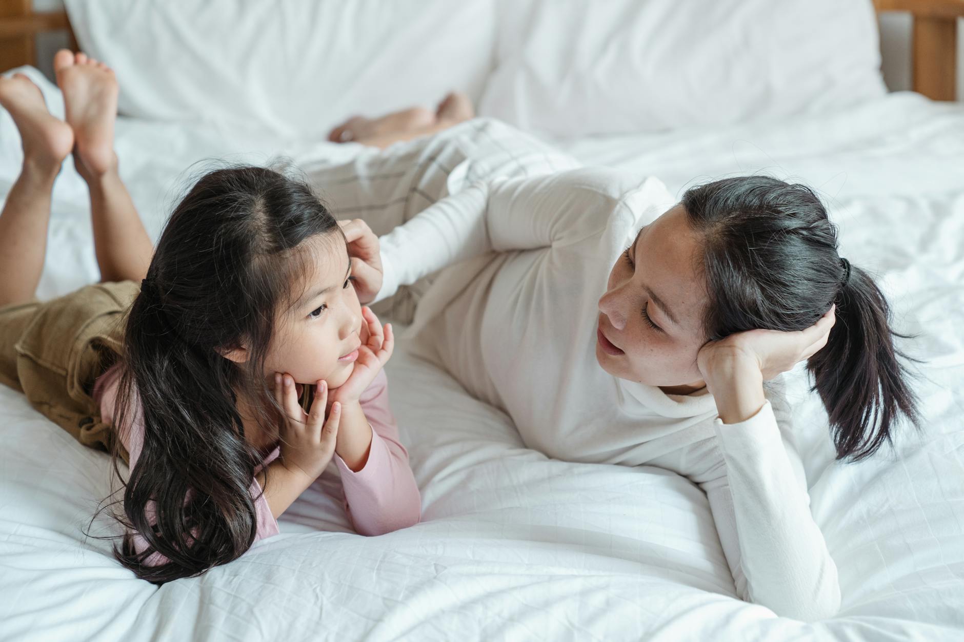 photo of woman and girl talking while lying on bed